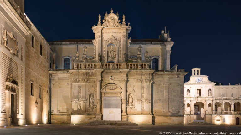 Illuminated Baroque church façade with carved portal and statues facing an empty stone square at night.