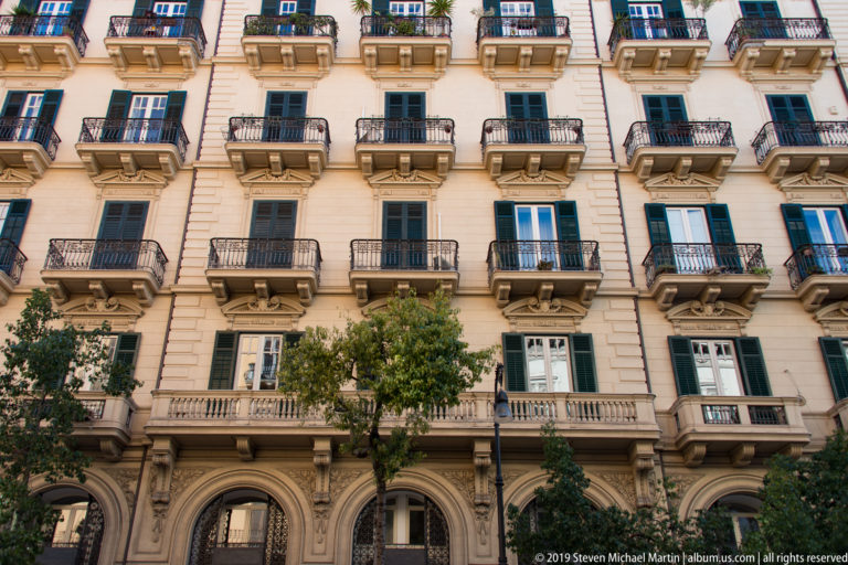 A pale Palermo apartment building has rows of wrought-iron balconies, planters and arched ground-floor openings.