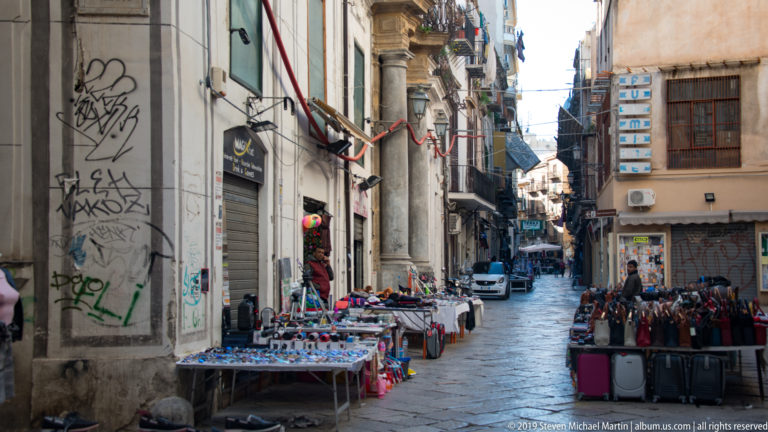 Tables and shopfronts line a narrow Palermo street beneath balconies and upper-story windows.