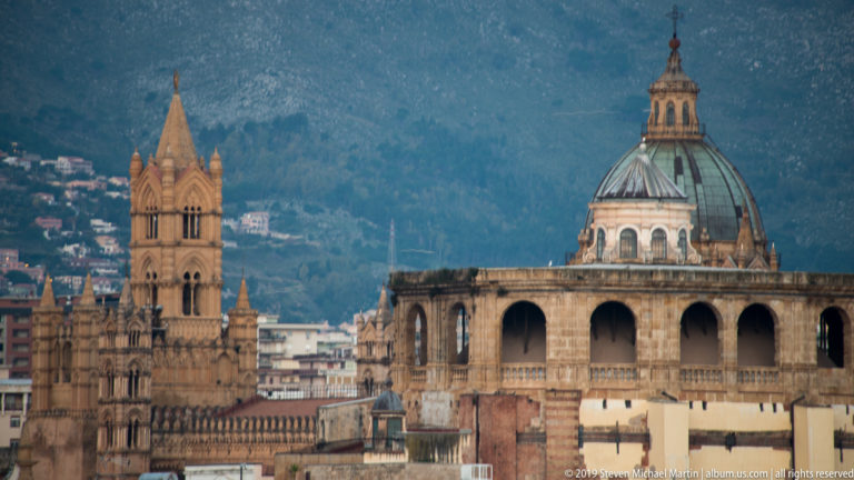 A pointed cathedral spire and a large church dome rise above Palermo’s rooftops at dusk.