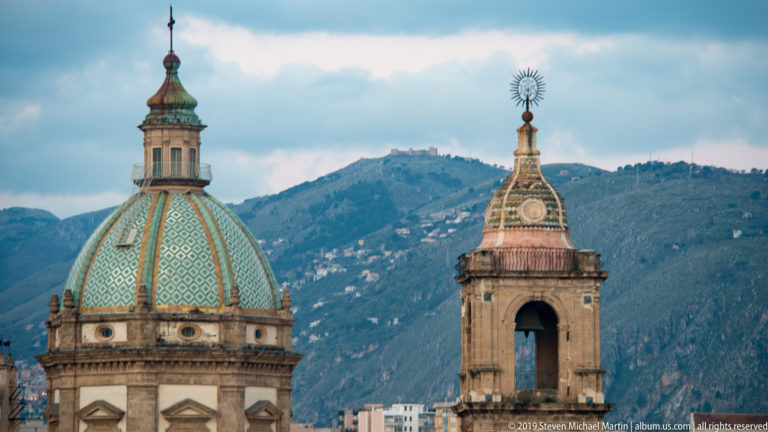 A patterned dome and an open bell tower stand side by side against Palermo’s hillside.