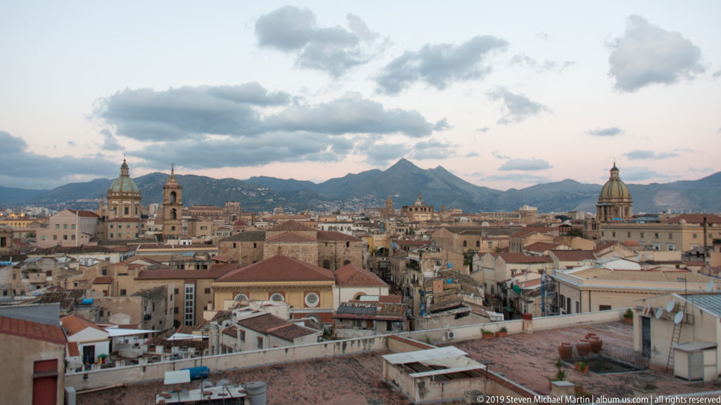 A rooftop terrace and layered Palermo roofs lead toward domes and towers beneath a cloudy evening sky.
