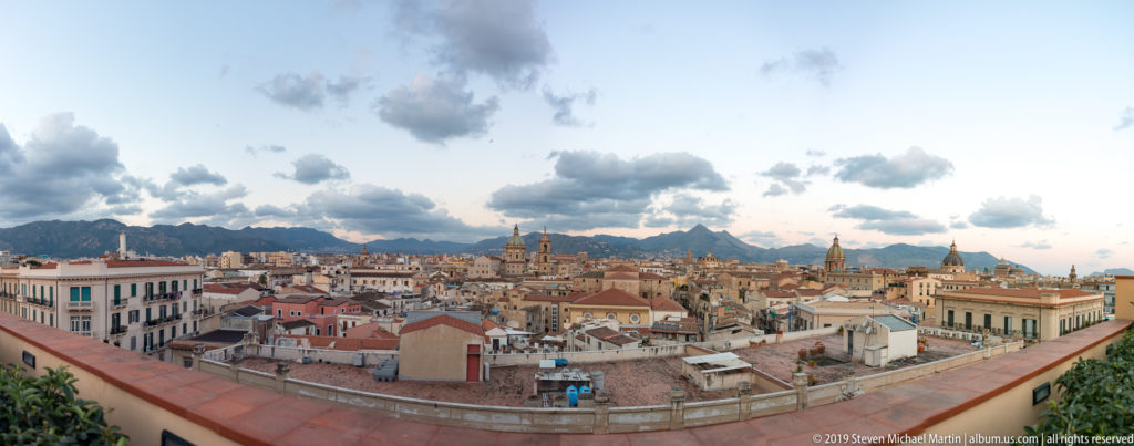 A rooftop terrace overlooks Palermo’s dense roofs, church towers, and Monte Pellegrino beneath open clouds.