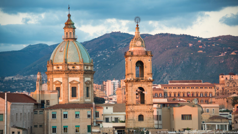 A green-patterned dome and open bell tower rise before Monte Pellegrino in Palermo.