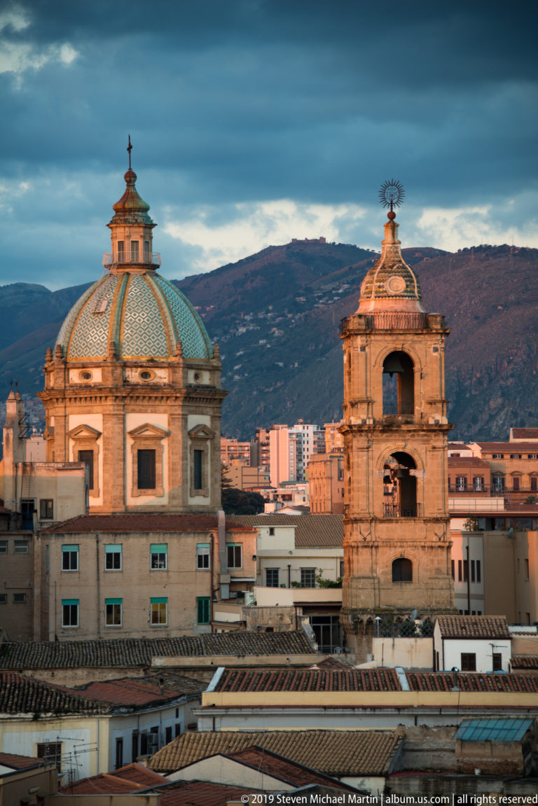 A patterned dome and a taller bell tower stand above Palermo’s rooftops and hillside backdrop.
