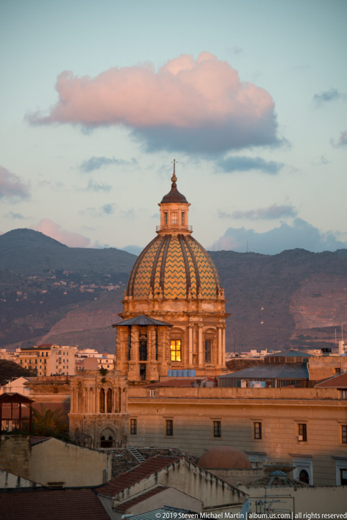 A warmly lit ribbed dome rises over shadowed Palermo rooftops beneath a pink-blue evening sky.