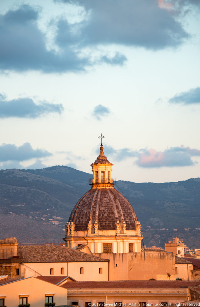 A patterned church dome and lantern glow above Palermo rooftops against a dark mountain ridge.