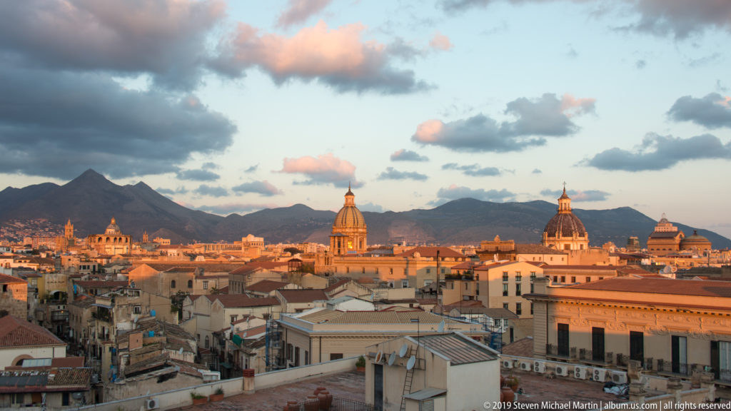 An elevated view crosses Palermo’s tiled roofs toward church domes and Monte Pellegrino at dusk.