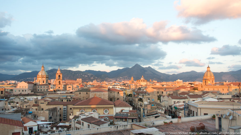 Palermo’s terracotta rooftops and church domes glow beneath clouds, with Monte Pellegrino on the horizon.