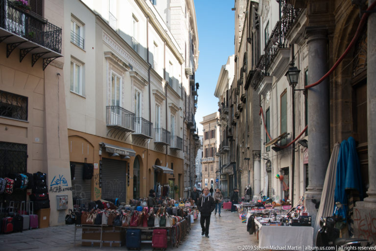 Shoppers walk through a narrow Palermo market lane lined with stalls and overhead signs.