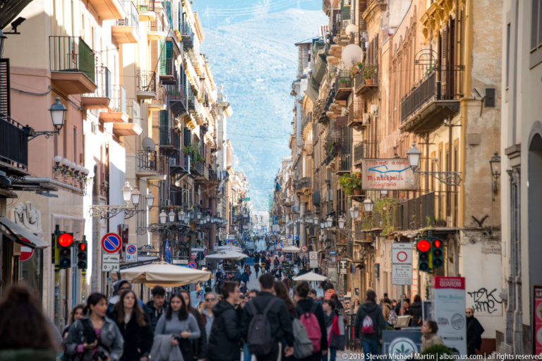 People cross a busy Palermo street lined with tall buildings, traffic signals and shop signs.