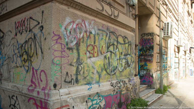 Colorful graffiti and overlapping tags cover a plaster wall beside a narrow Palermo street.
