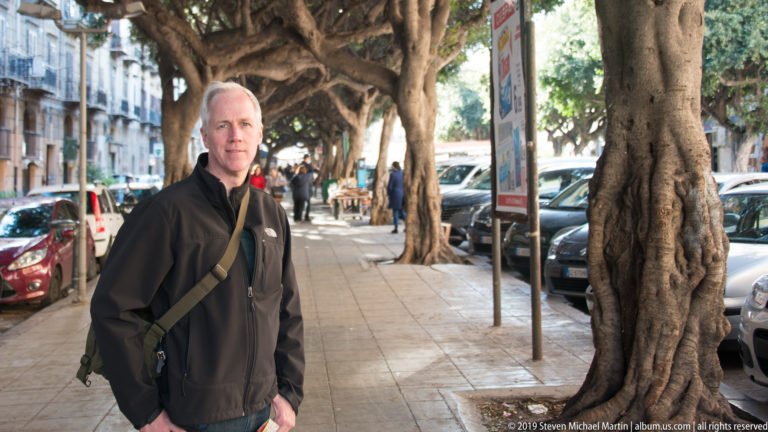 A man in a dark jacket stands beneath trees beside parked cars and pedestrians in Palermo.