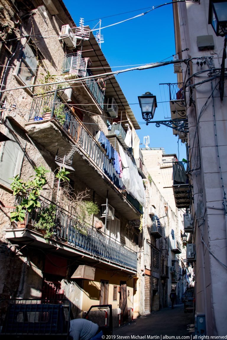 A narrow Palermo street is lined with tall buildings, balconies, awnings and shopfronts.