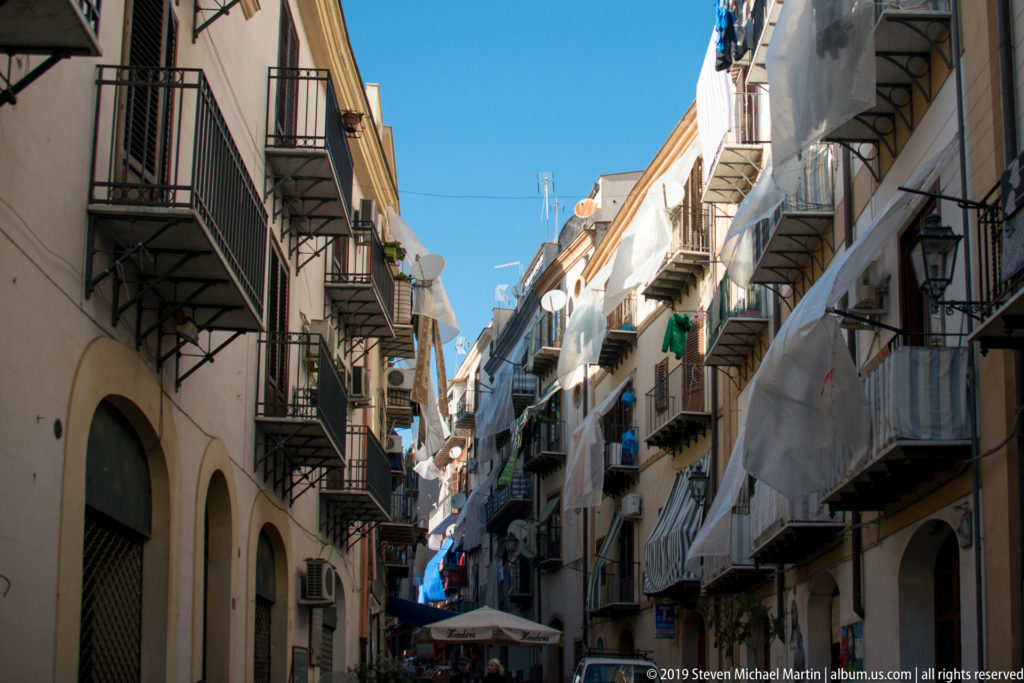 A shaded Palermo lane runs between close-set homes with balconies and shuttered windows.