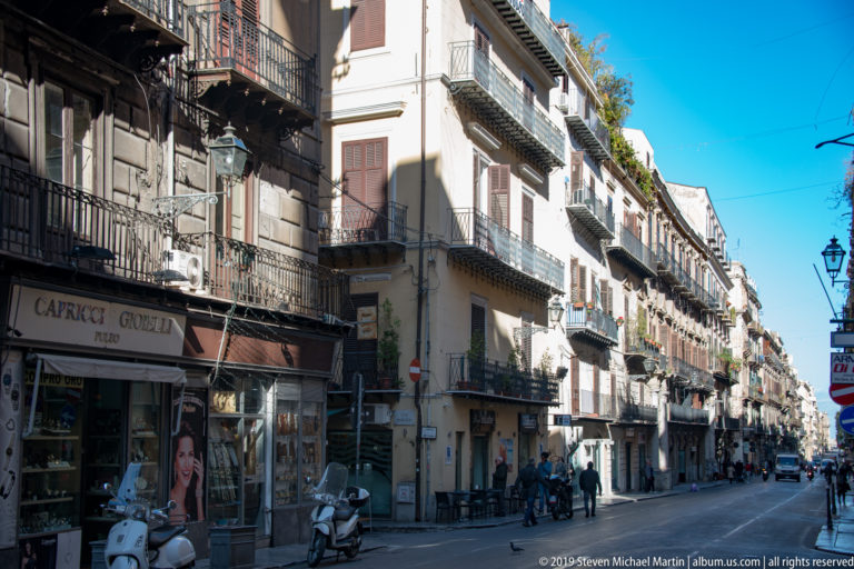 A Palermo avenue lined with balconies, shop signs and storefronts recedes toward a bright distance.