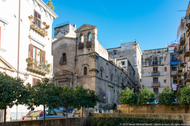 A small stone church with an arched tower stands among taller Palermo buildings and trees.