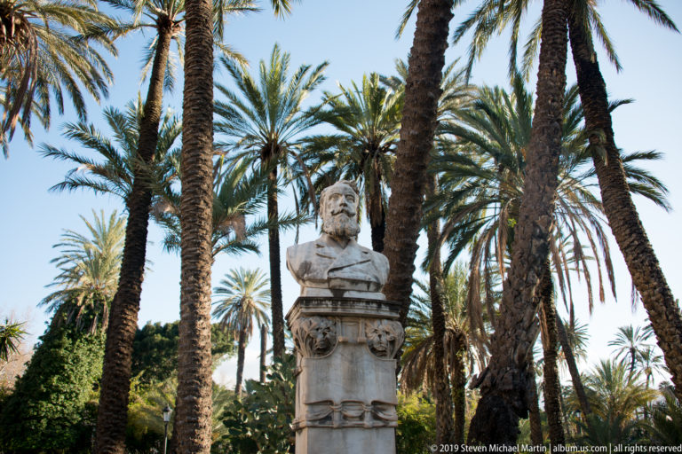 A stone bust on a tall pedestal stands among palms in a Palermo garden.
