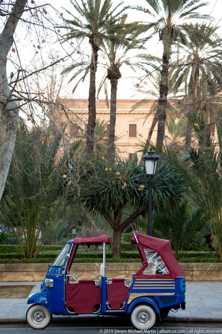 A red-and-blue three-wheeled taxi drives past palm trees on a Palermo street.