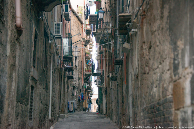 A narrow alley runs between tall Palermo buildings, with small figures visible in the distance.
