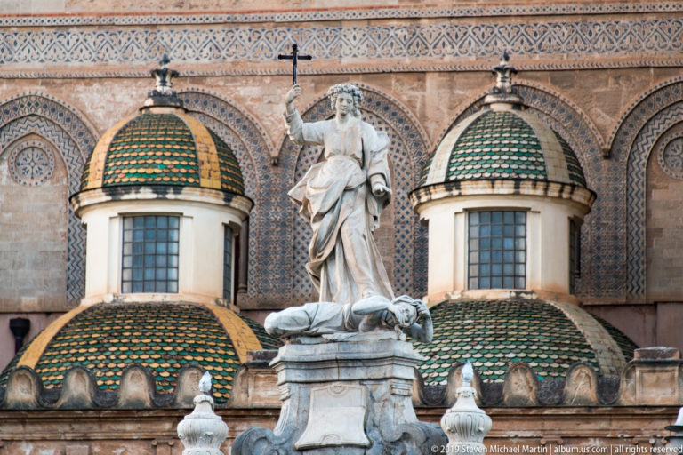 A pale standing statue rises before the green-and-gold tiled dome and patterned façade of Santa Caterina.