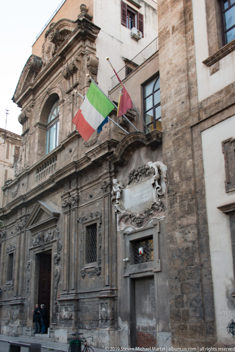 Italian flags hang above a doorway and balcony on a pale Palermo building façade.
