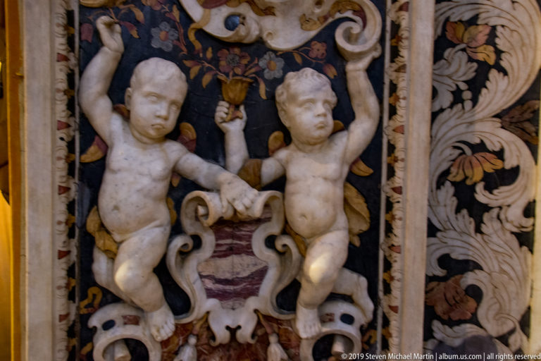 Two carved cherubs support an ornament within a framed stone relief panel.