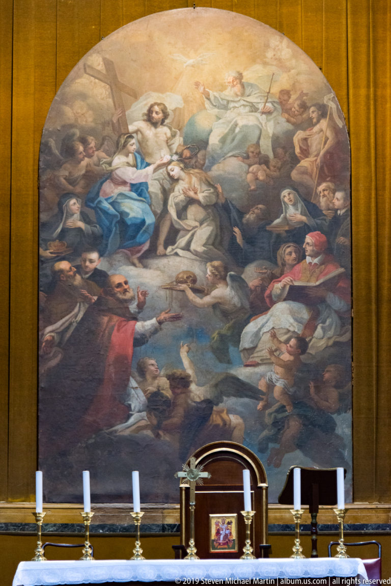 A crowded religious painting rises above an altar and candleholders inside a Palermo church.