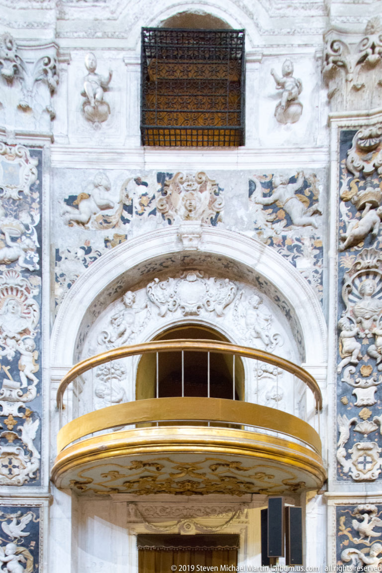 A curved balcony rail overlooks an ornate church interior with arches and painted decoration.