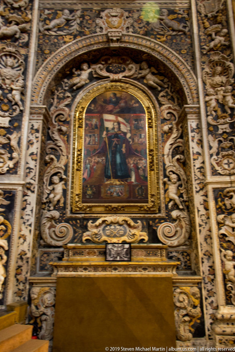 A large religious painting is enclosed by a carved gilded frame above a church altar.