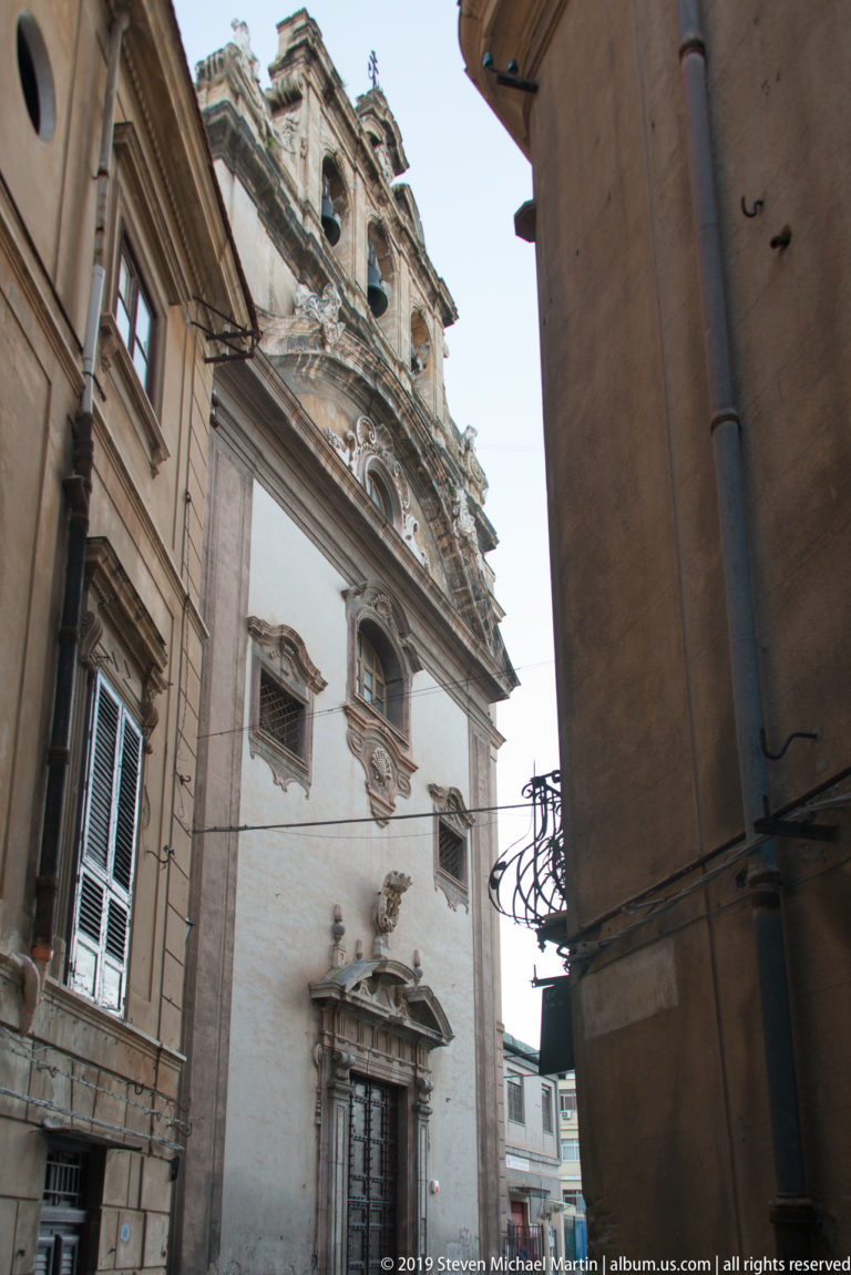 A pale multi-story building with balconies and a carved portal stands at a Palermo street corner.