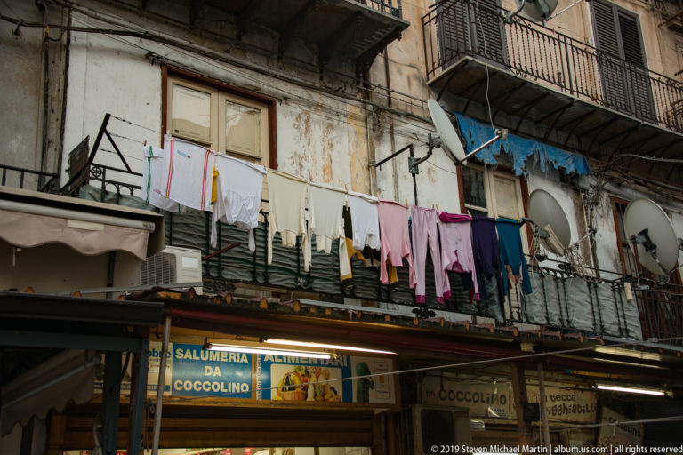 Laundry hangs above a salumeria sign and awning on a narrow Palermo street.
