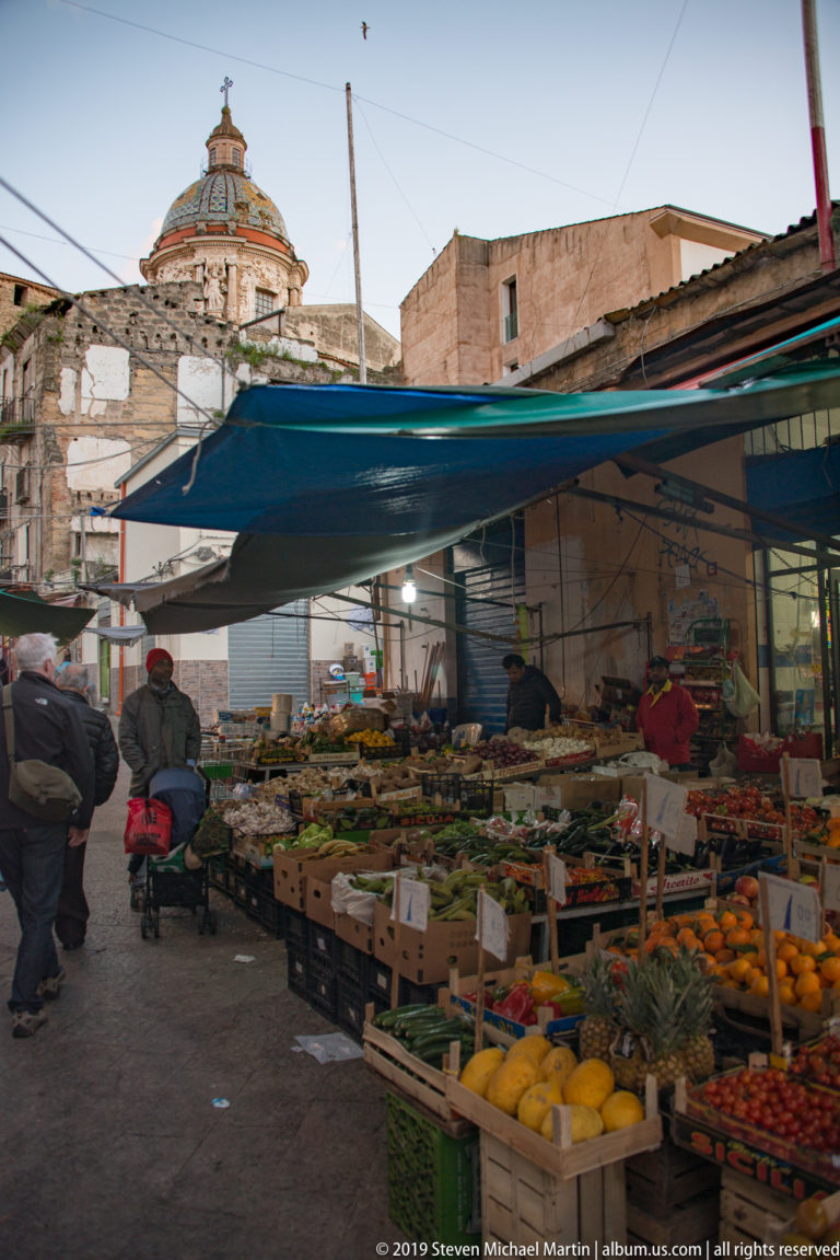 A Palermo produce stall displays citrus and other goods beneath blue awnings along a narrow street.