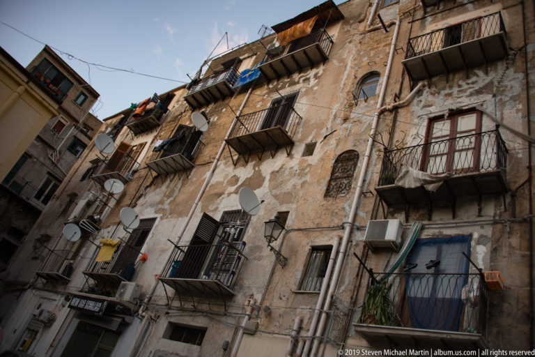 A narrow Palermo lane is enclosed by tall homes with iron balconies and projecting awnings.
