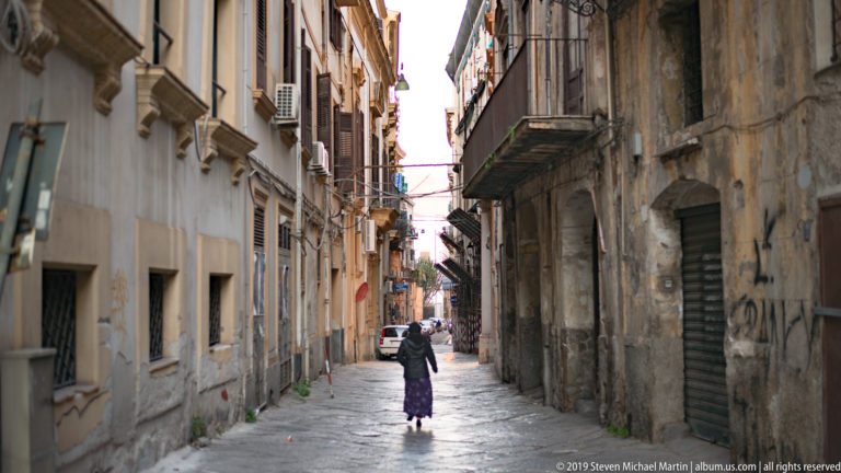 A pedestrian walks away along a narrow street lined with tall façades and balconies.
