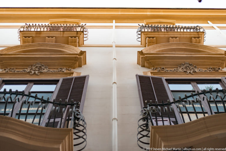 An upward view of a Palermo façade with iron balconies, dark shutters and projecting wooden eaves.