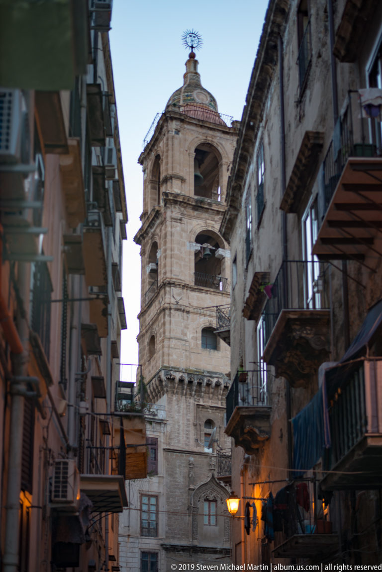 A narrow street of tall façades and balconies leads toward a domed church in Palermo.