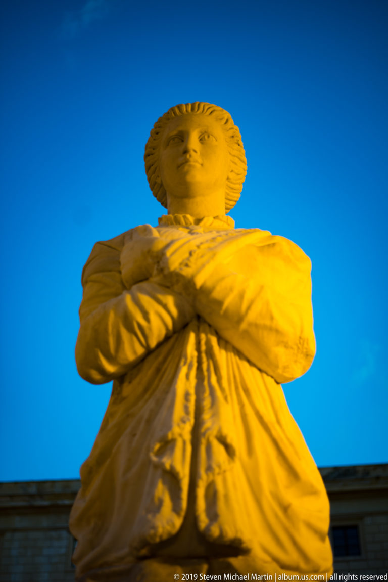 A marble male figure with folded arms stands above the fountain rim against blue sky.