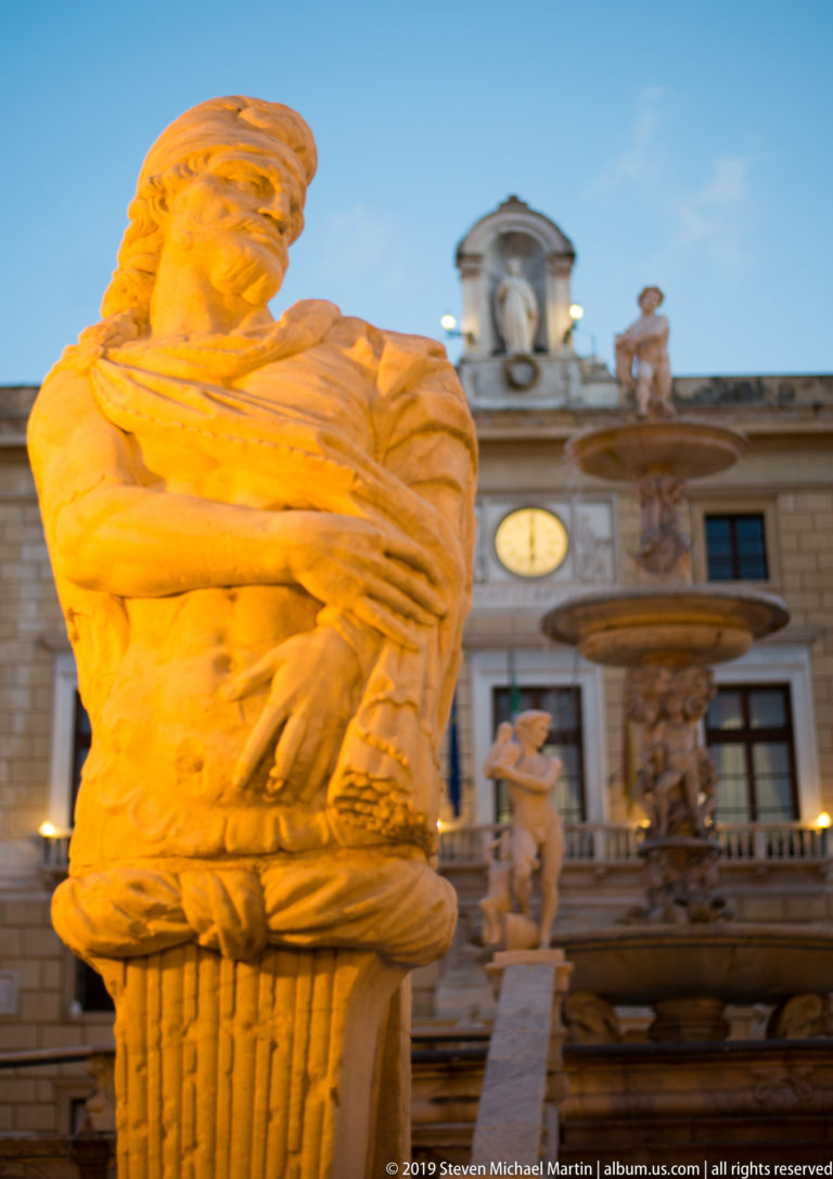 A pale stone figure crowns a fountain in front of a historic Palermo façade at dusk.