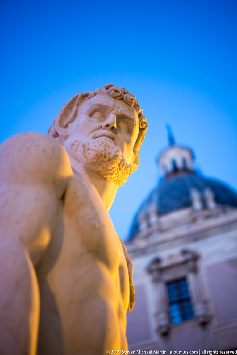 Close view of a carved male head and torso at Fontana Pretoria, with another statue and dome behind.