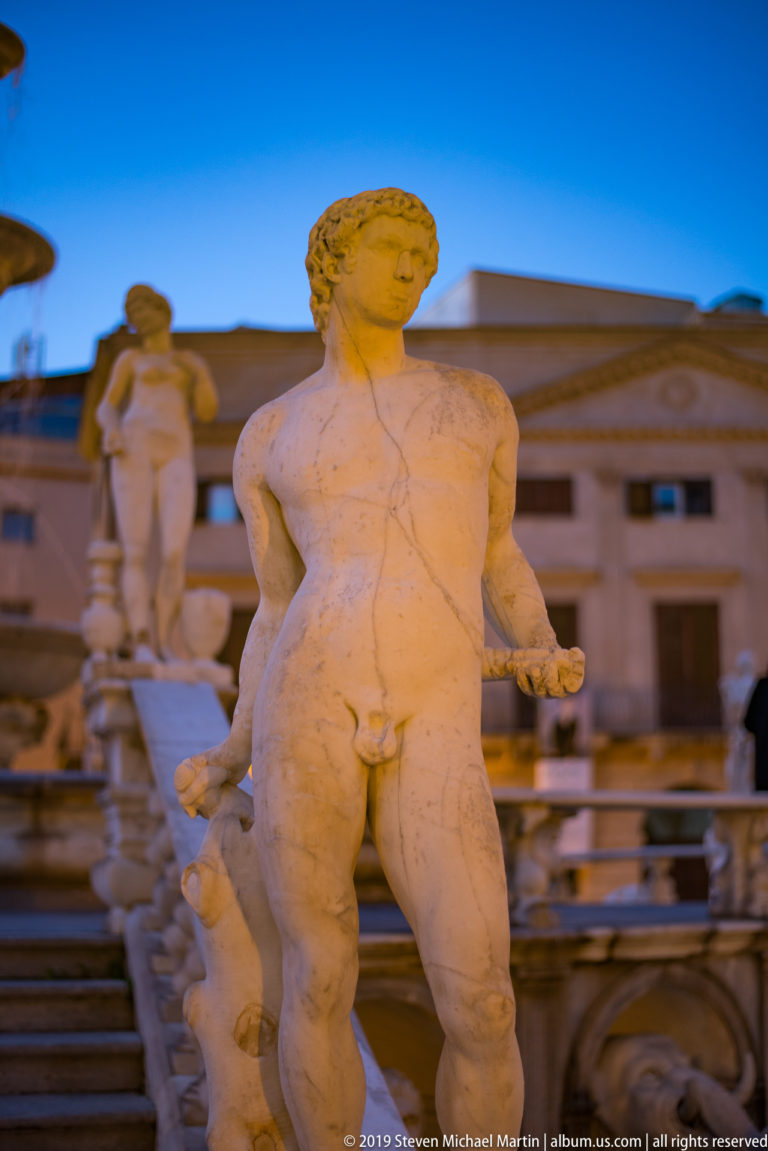 A nude marble figure gestures outward from Fontana Pretoria against a Palermo façade.