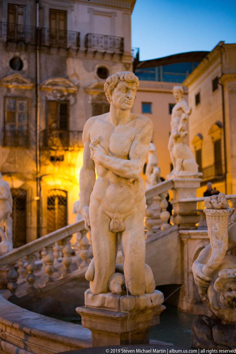 A standing marble male figure rises above the fountain as neighboring statues and façades recede behind.