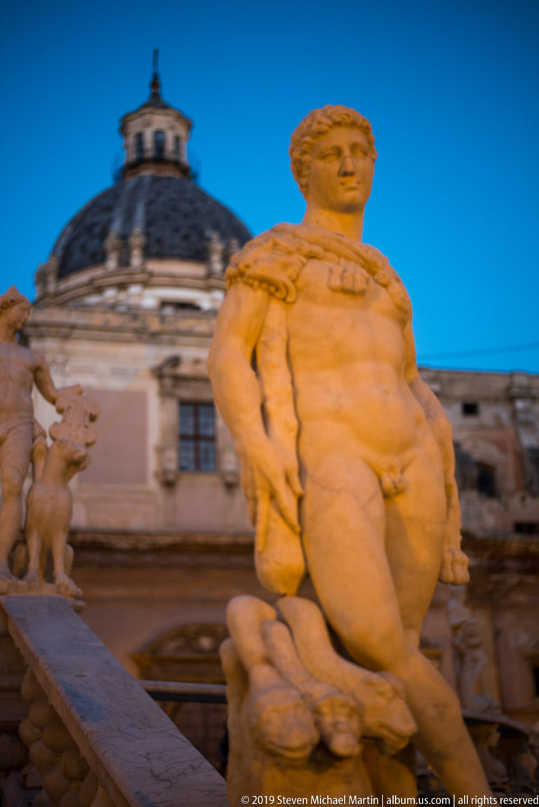 A standing marble figure on Fontana Pretoria is framed against a dark church dome and blue sky.