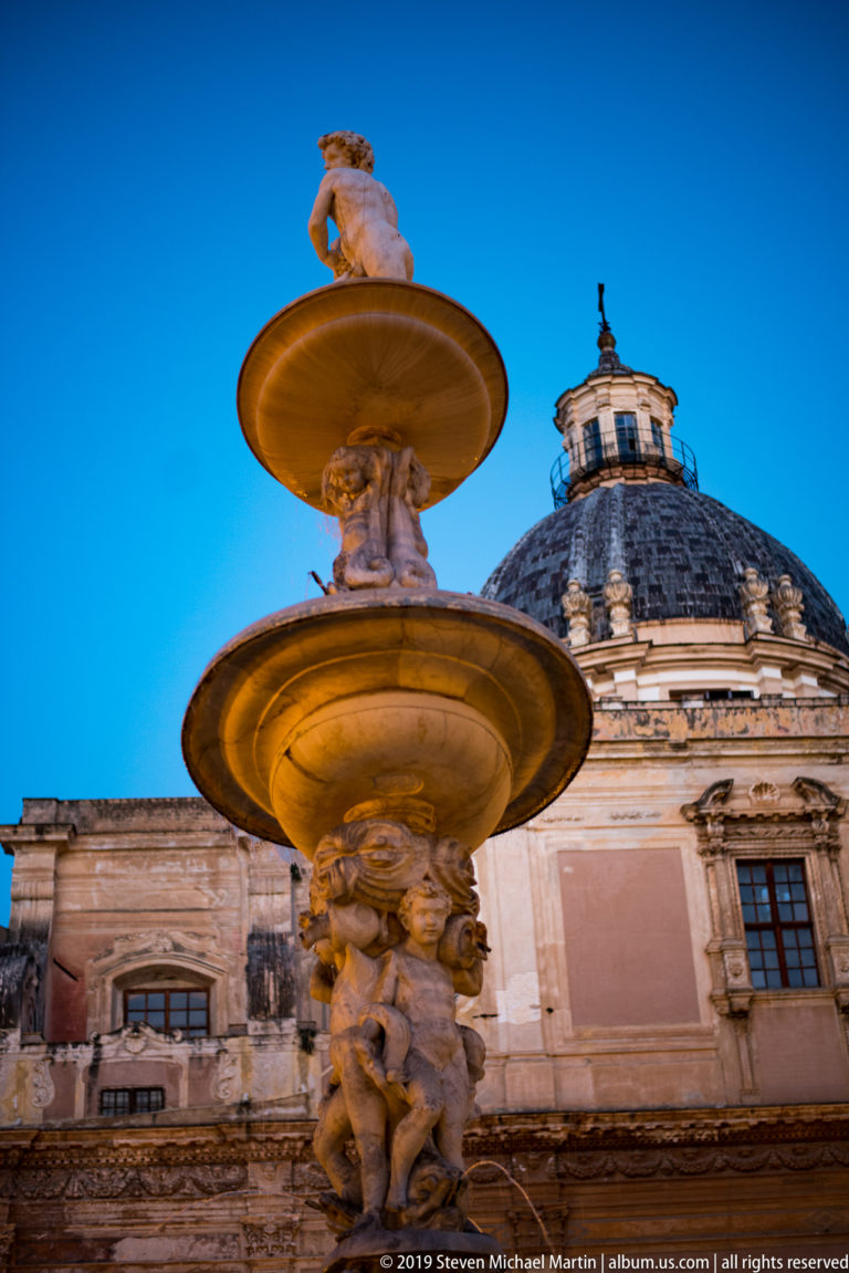 A marble figure rises above the lower basin of Fontana Pretoria, with Palermo buildings behind it.