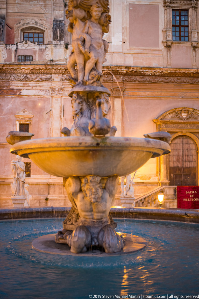A tiered stone fountain with a broad basin and carved figures stands beside a Palermo street.