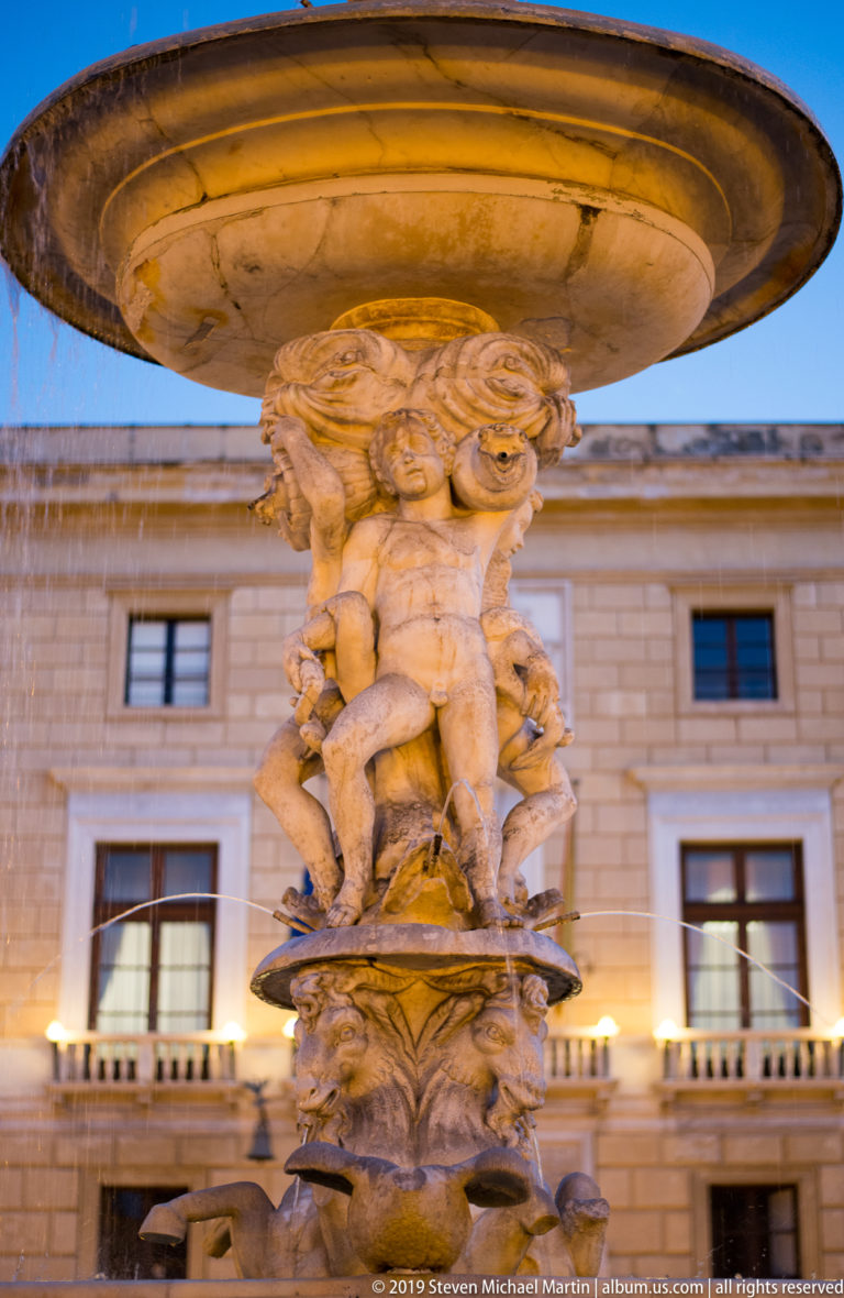 Carved stone figures support a broad fountain basin against a pale Palermo façade.