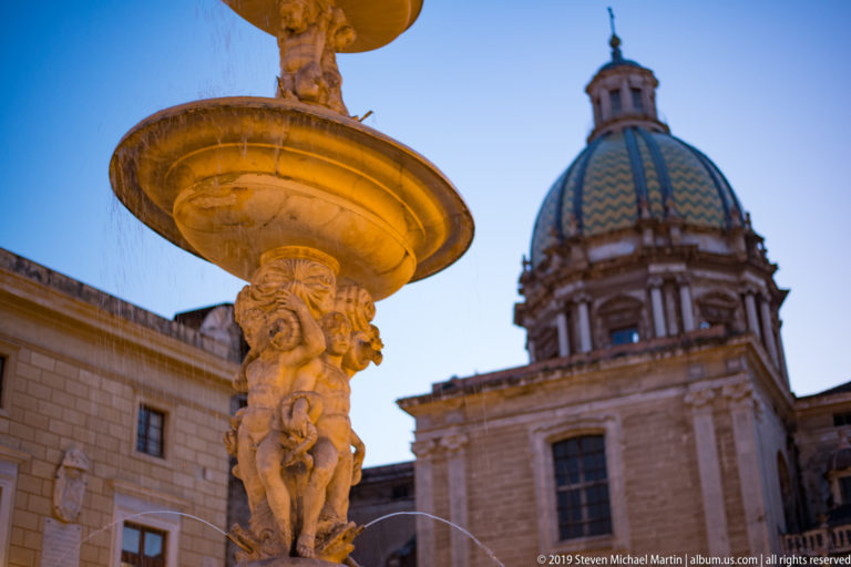 A stone figure supports a fountain basin before a patterned church dome at dusk.