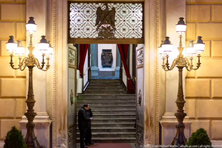 Twin lamps frame a stairway and dark entrance beneath a patterned panel in a Palermo building.