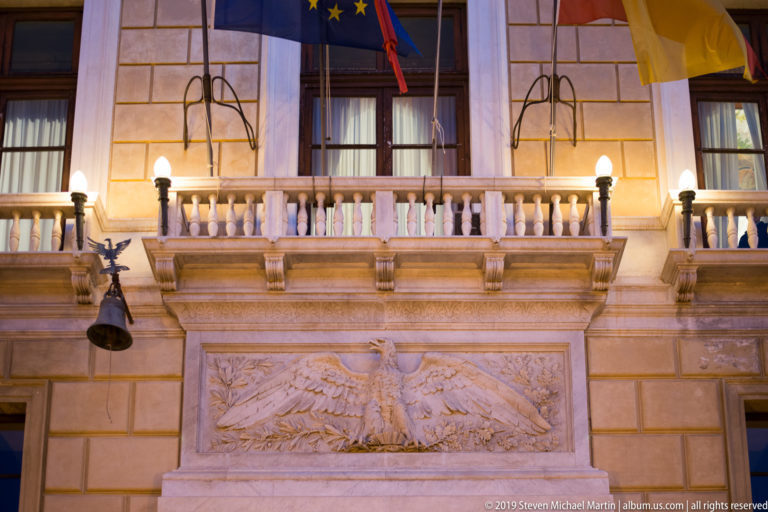 An ornate interior pairs a balustraded balcony with banners and a large sculpted wall panel.