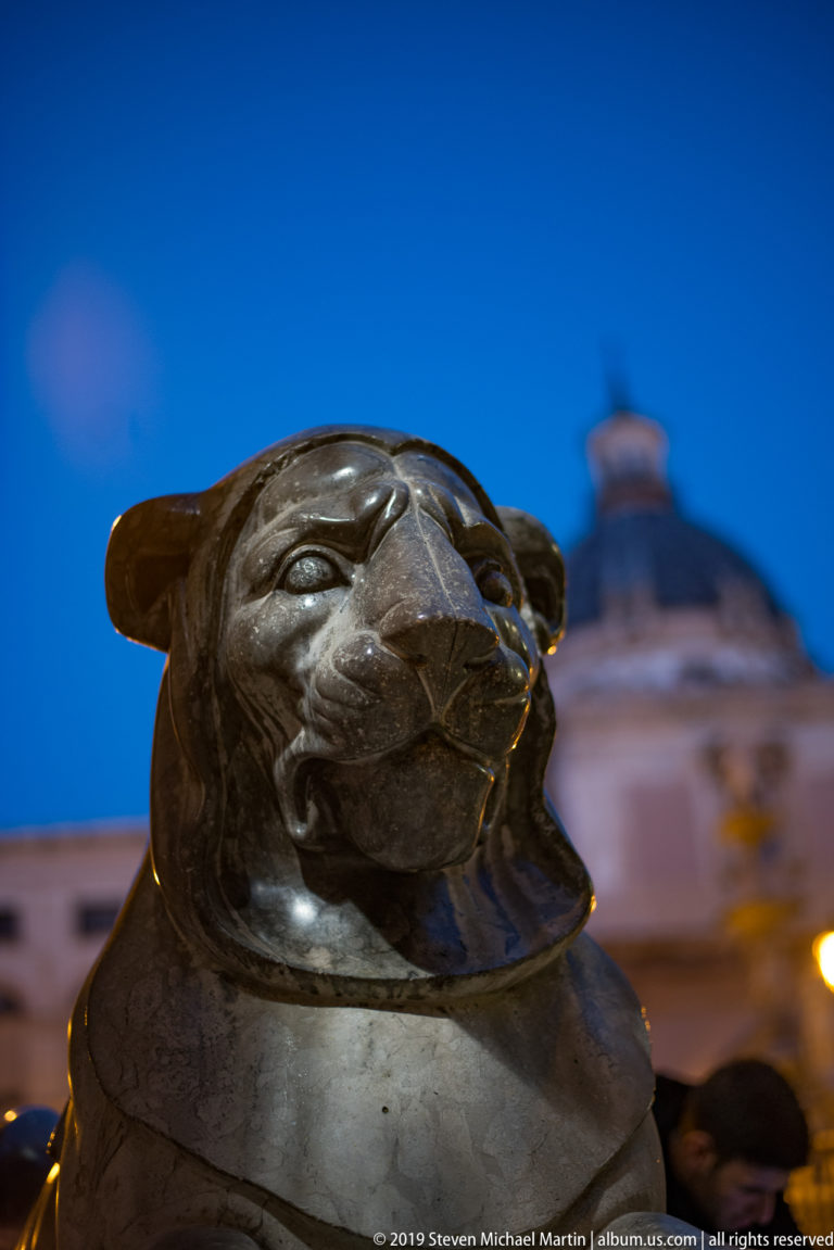 A weathered stone head with closed eyes is shown in close-up against a blurred dusk city.